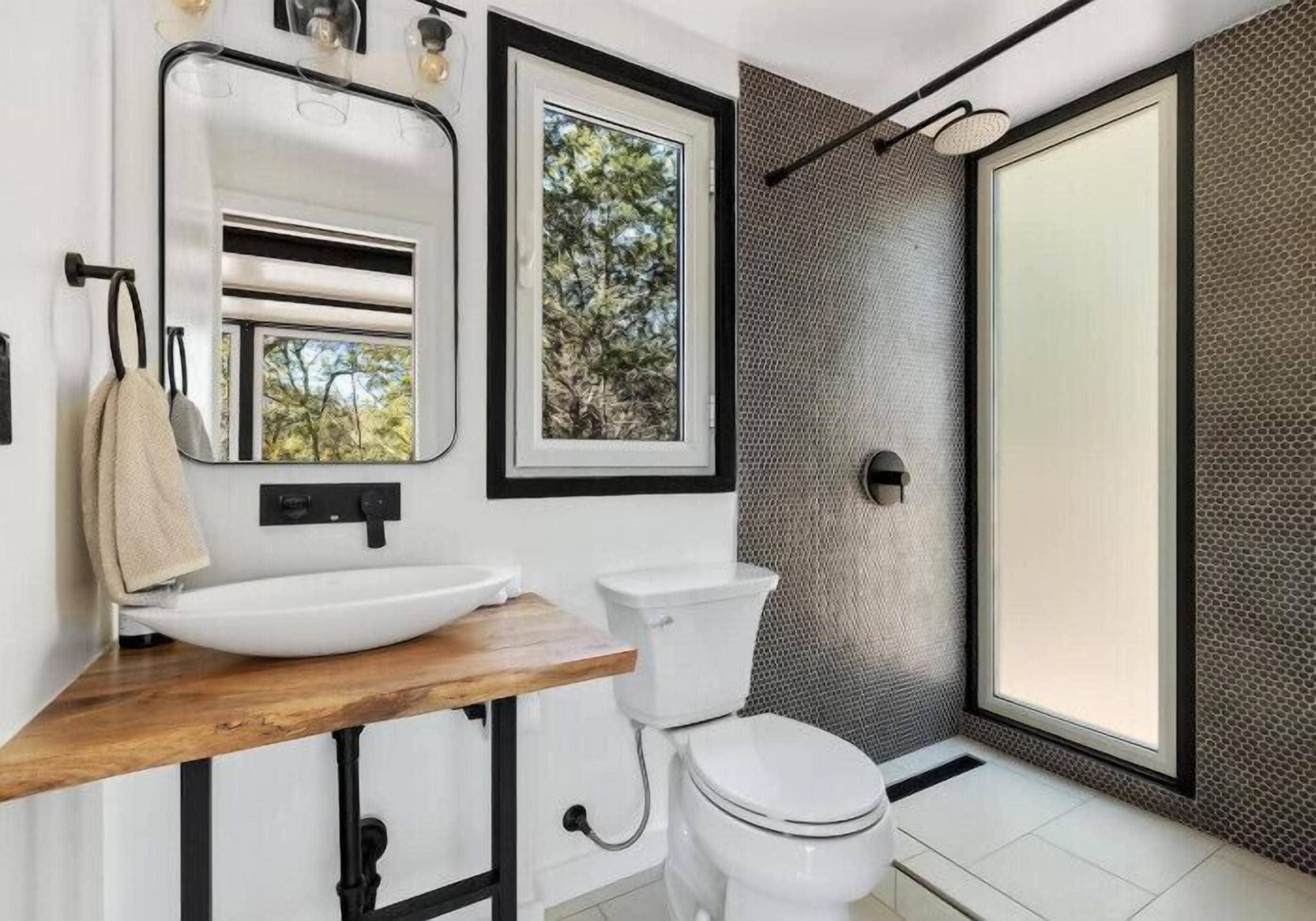 A modern bathroom with a wooden vanity, a white toilet, a large mirror, and a shower with a textured wall.