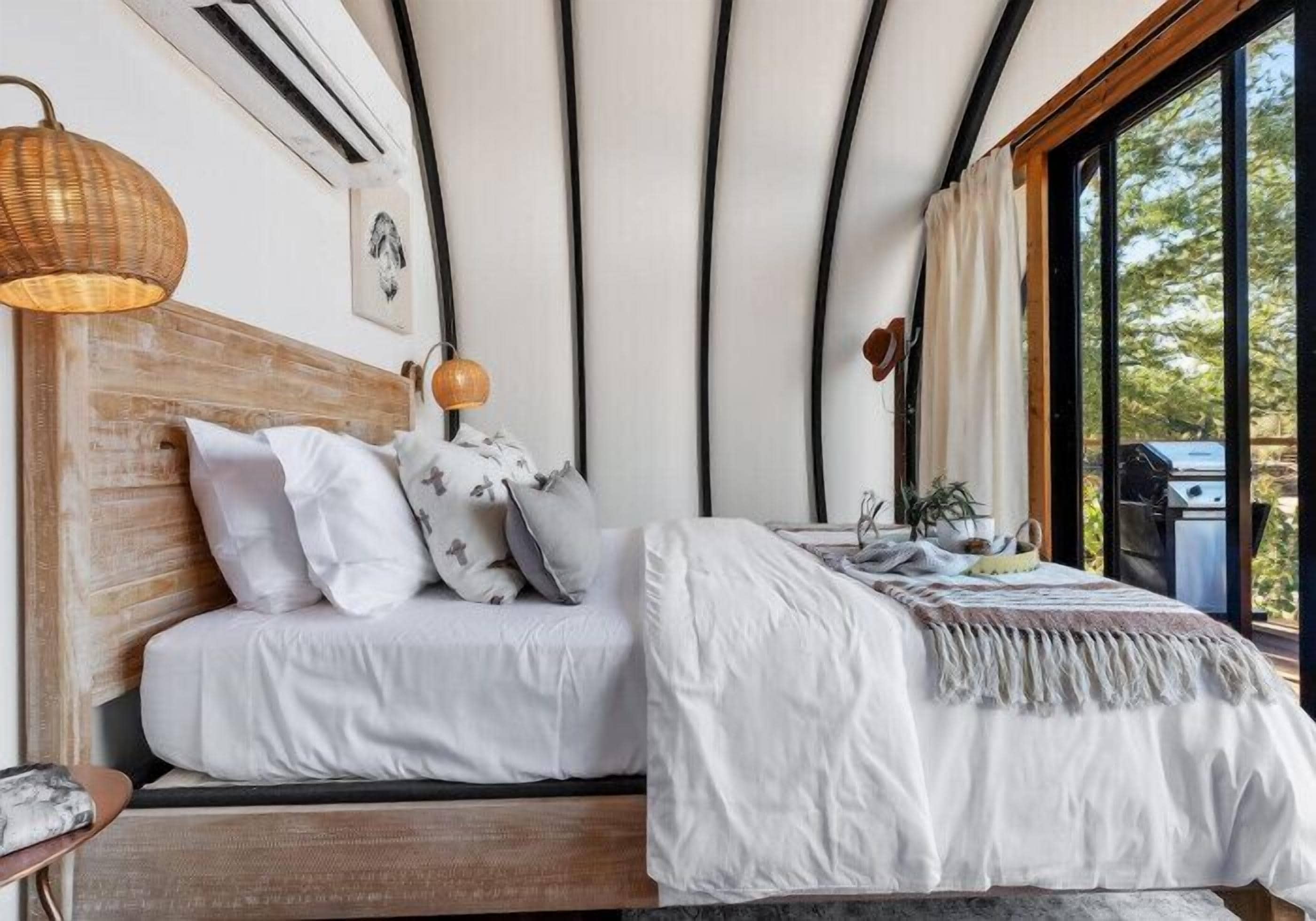 Ein Schlafzimmer mit einem Holzkopfende, weißer Bettwäsche, einer Nachttischlampe und Blick ins Freie durch große Fenster.