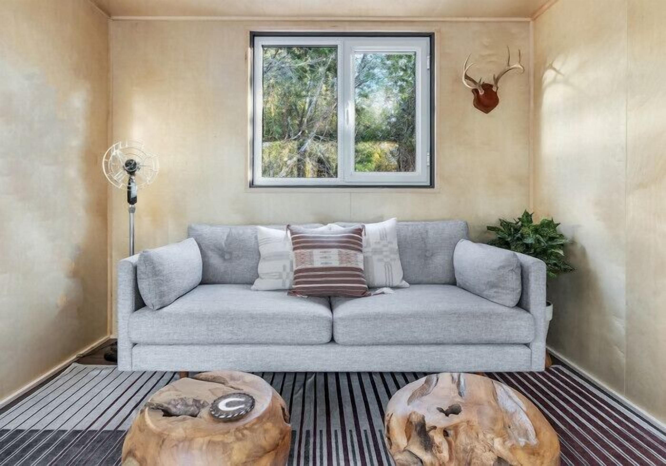 A room with a grey sofa, a wooden coffee table, a floor lamp, a window with a view of trees, and a mounted deer head on the wall.