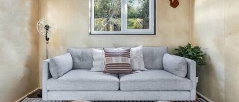 A room with a grey sofa, a wooden coffee table, a floor lamp, a window with a view of trees, and a mounted deer head on the wall.