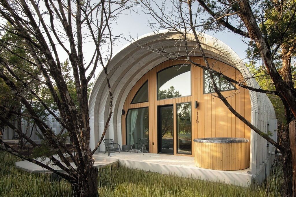 A modern, circular building with large windows and a wooden exterior, surrounded by trees and a grassy area.