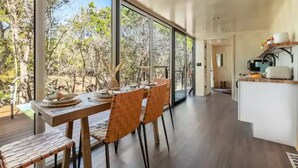 Un espace repas moderne avec une table et des chaises en bois, de grandes fenêtres donnant sur la verdure et un coin cuisine doté de meubles et d’appareils électroménagers blancs.