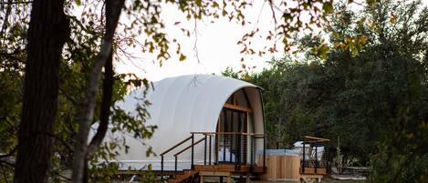 A dome-shaped cabin with a wooden deck and a small wooden structure adjacent to it, surrounded by trees.