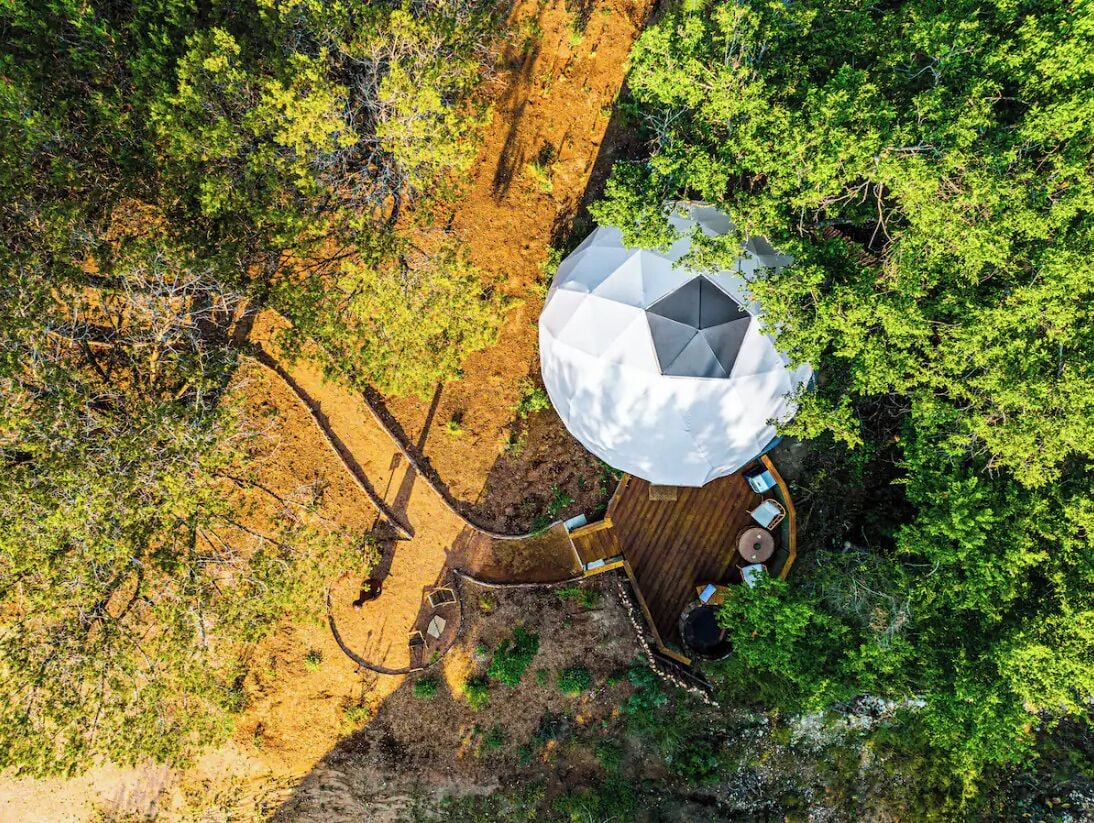 Une vue aérienne d’une structure moderne en forme de dôme, dotée d’une terrasse en bois et d’un espace salon, le tout entouré d’une végétation luxuriante.