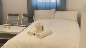 Comfortable room with a neatly made bed, a table, and a chair, featuring a window with a curtain and a control panel.