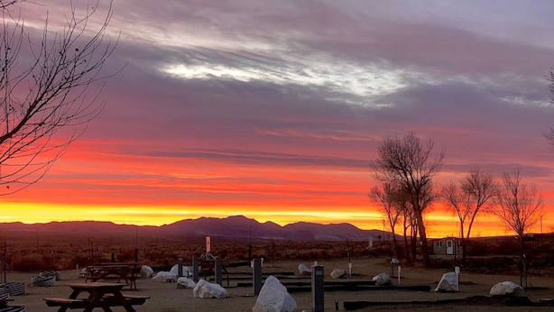 Un terrain de camping situé dans le désert, équipé de tables de pique-nique, de rochers et offrant une vue dégagée sur les montagnes au coucher du soleil.