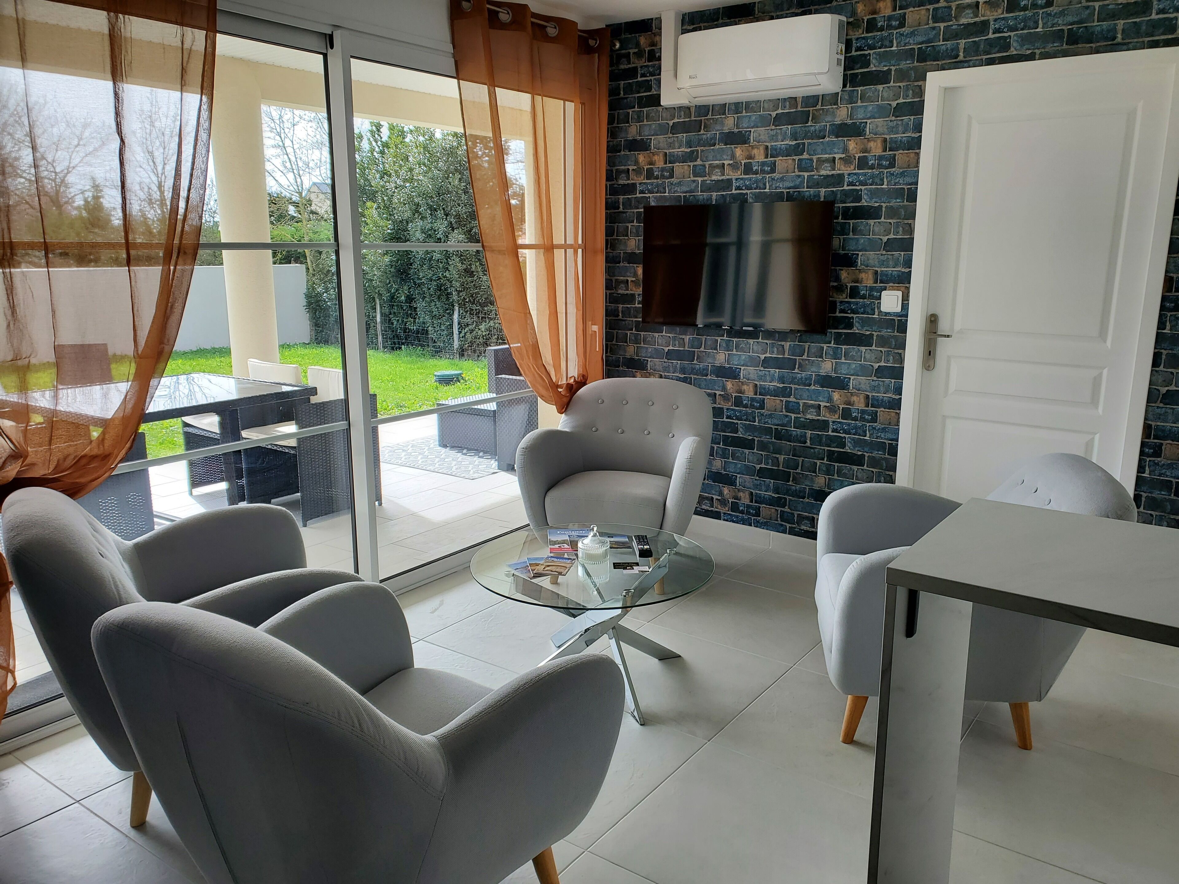 Un salon moderne avec une table basse en verre, des fauteuils gris et un téléviseur blanc fixé au mur en briques.