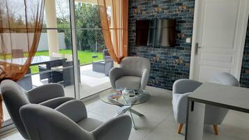 Un salon moderne avec une table basse en verre, des fauteuils gris et un téléviseur blanc fixé au mur en briques.