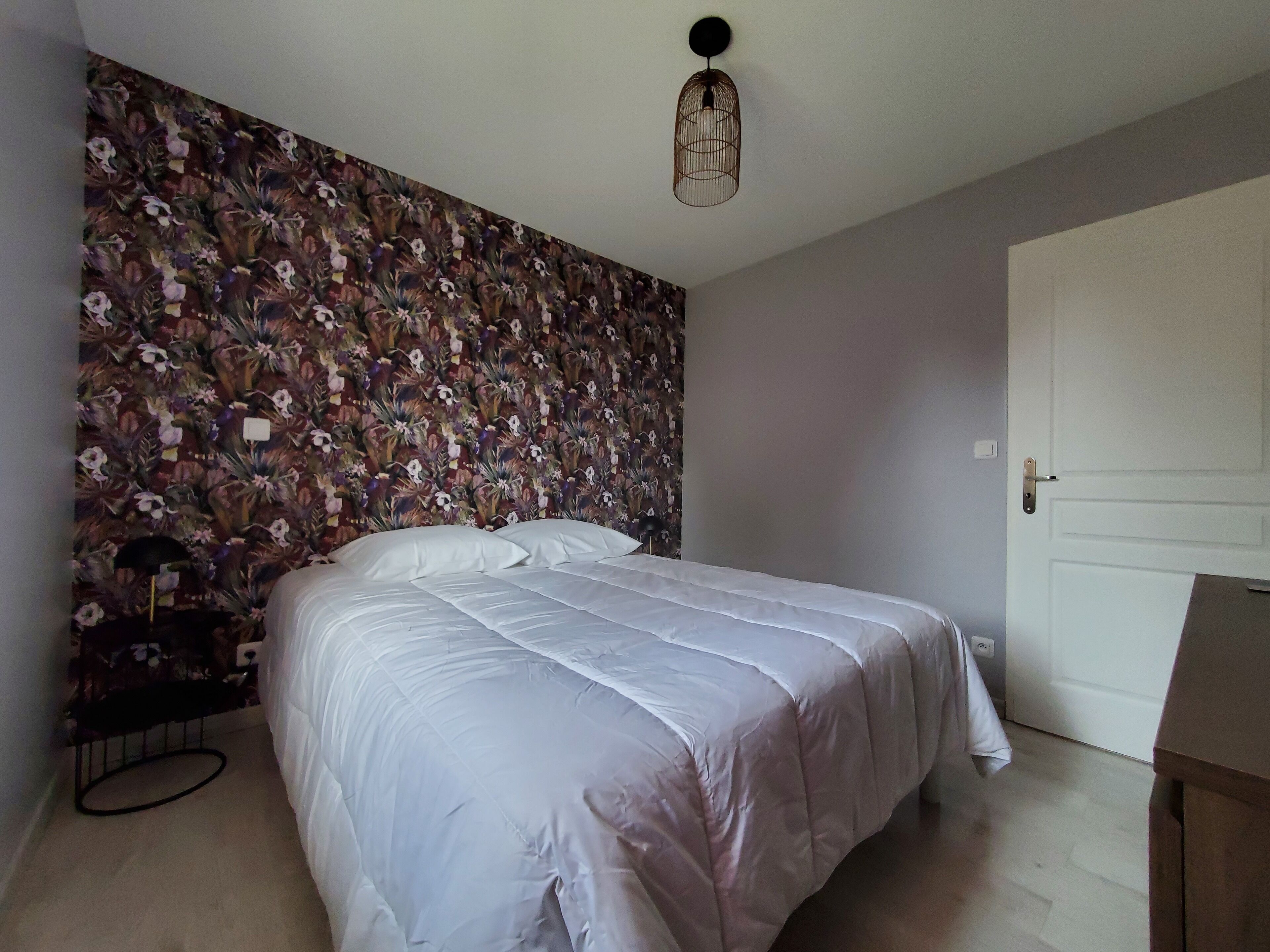 A bedroom with a floral wallpaper, a bed with white bedding, a hanging light fixture, and a door.