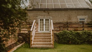 A rustic building with a wooden staircase leading to a white door, surrounded by greenery and a wooden fence. - Four Season's Splendor: Artist's Nest beside Walled Garden on Seaside Estate (Portpatrick)