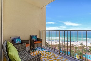 Un balcón con mobiliario de mimbre y un tapete estampado, con vistas a una playa con sombrillas y una piscina.