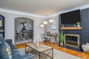 Ein modernes Wohnzimmer mit Sofa, Couchtisch und Kamin. Es gibt einen Essbereich mit Stühlen und Tisch, und im Hintergrund eine Küche.