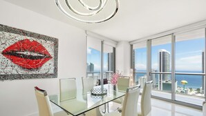 Uma sala de jantar moderna com mesa e cadeiras de vidro, um grande quadro abstrato, e vista para a cidade e o oceano.