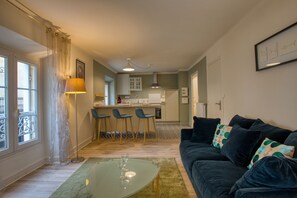 A modern living room with a dark sofa, a glass coffee table, and a kitchen in the background.