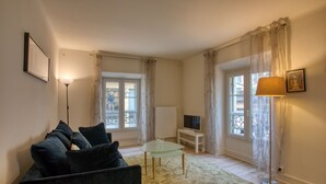 A living room with a sofa, a glass coffee table, a TV stand, and a lamp. - Appartement T3 de 60 m2 - Centre Historique (Sarlat-la-Canéda)