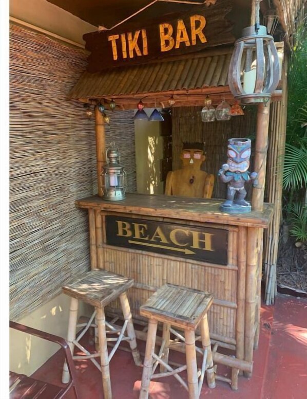 A tiki bar with a sign reading "TIKI BAR" and "BEACH" indicating its location. There are two stools in front of the bar. - Captin' Joe's Tropical Palm Beach Hot Tub Hideaway (West Palm Beach)