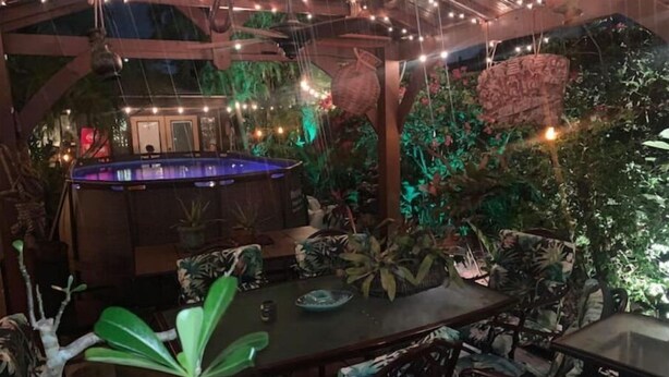 An indoor pool area with a table and chairs, surrounded by tropical plants and decorative lighting.