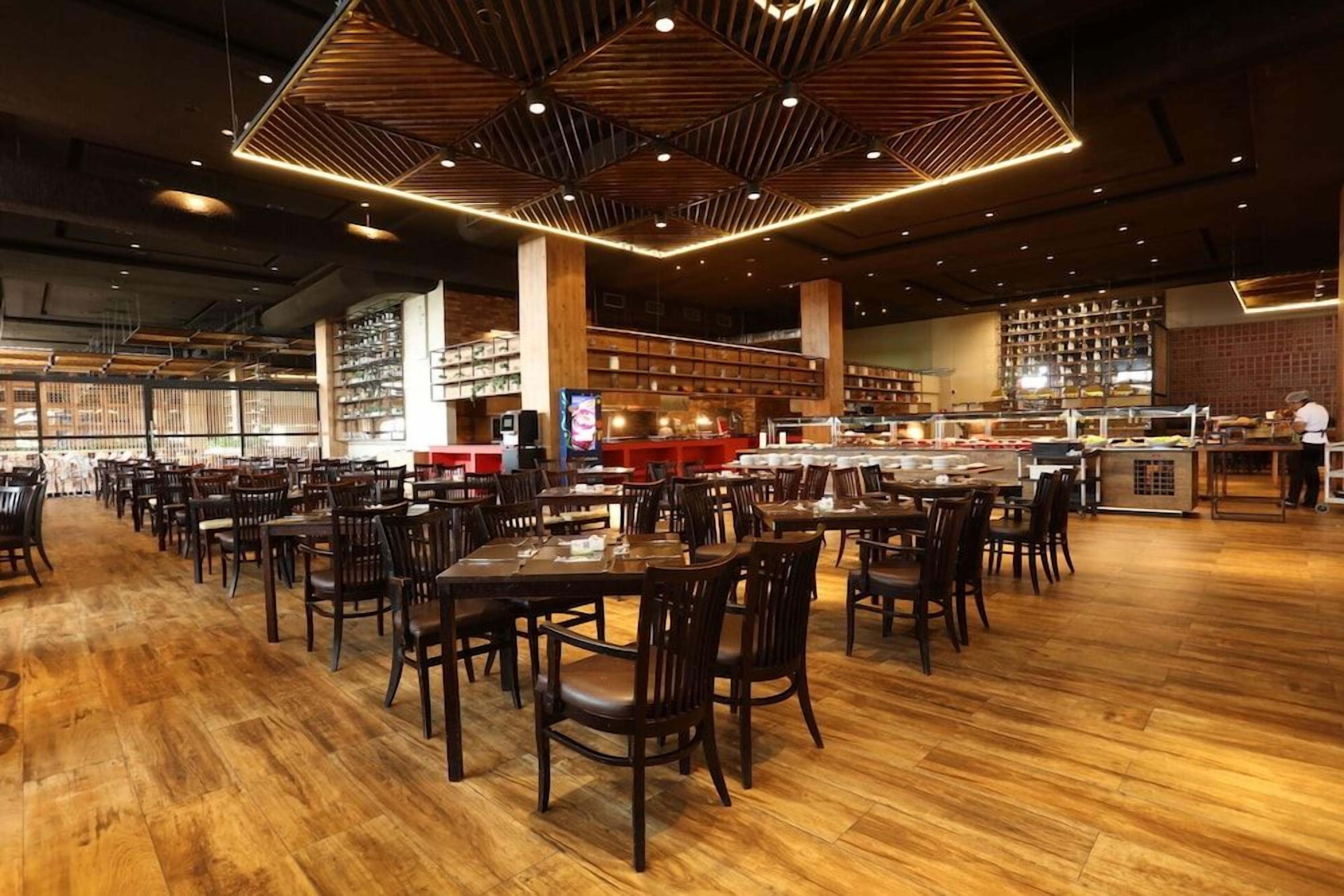 A spacious restaurant with multiple dining tables and chairs, a kitchen area, and a variety of shelves in the background.