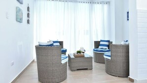 Een moderne woonkamer met rieten meubels, een glazen bijzettafeltje en uitzicht op het groen door dunne gordijnen.