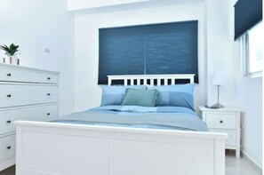 Une chambre moderne avec un lit blanc, une tête de lit bleue et une fenêtre avec des stores.
