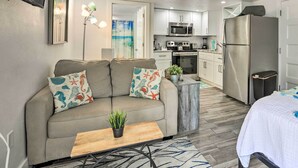 Een modern appartement met een grijze bank, een klein houten tafeltje, een keuken met witte kasten, een roestvrijstalen koelkast en een tv die aan de muur is bevestigd.