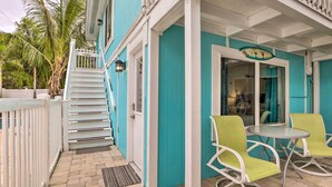 Een turquoise gebouw met een witte trap naar een deur, een klein buitentafeltje met stoelen en een palmboom op de achtergrond.