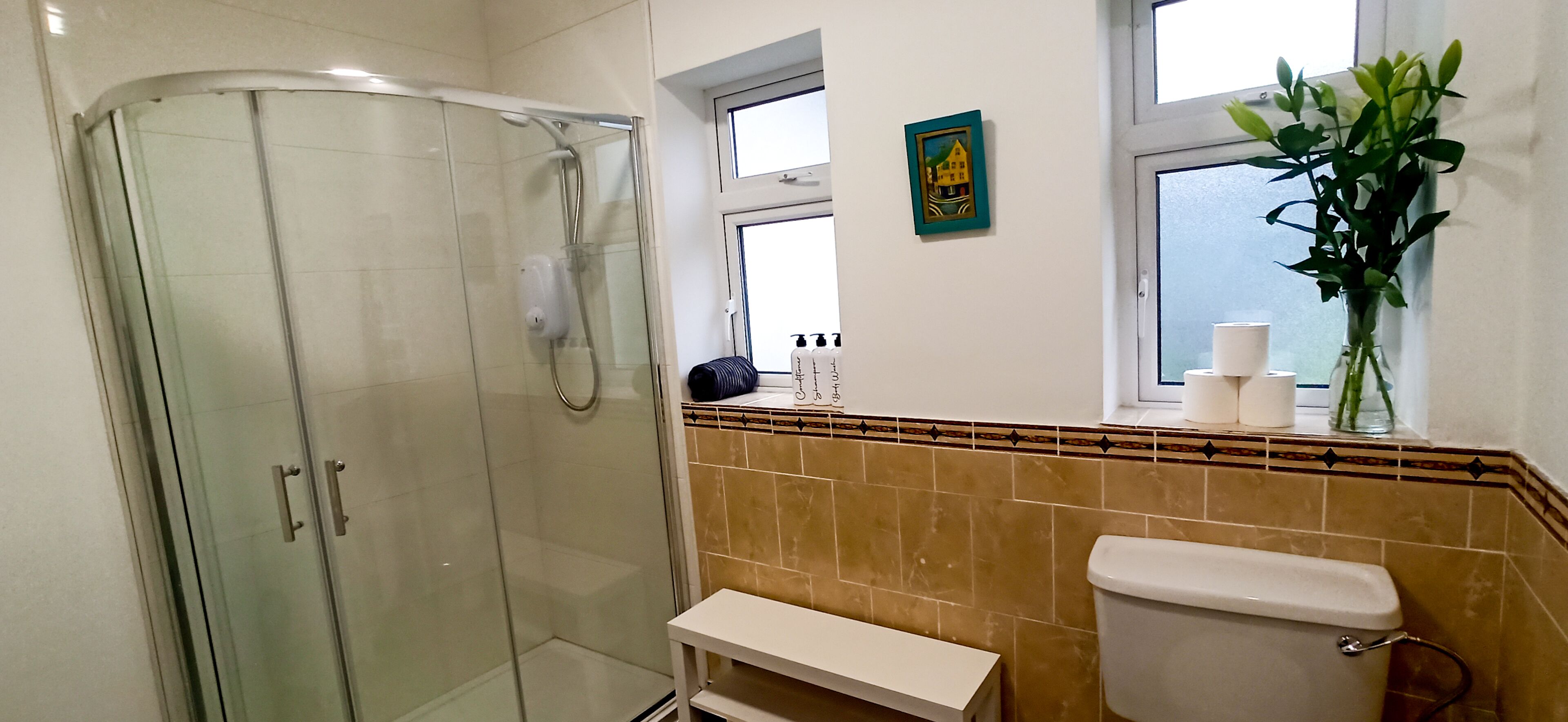 Ett badrum med en duschkabin i glas, en toalett, ett handfat med hygienartiklar och ett fönster med en växt.
