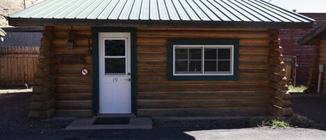 A small log cabin with a green metal roof, a white door with the number 19, and a window with a green frame.