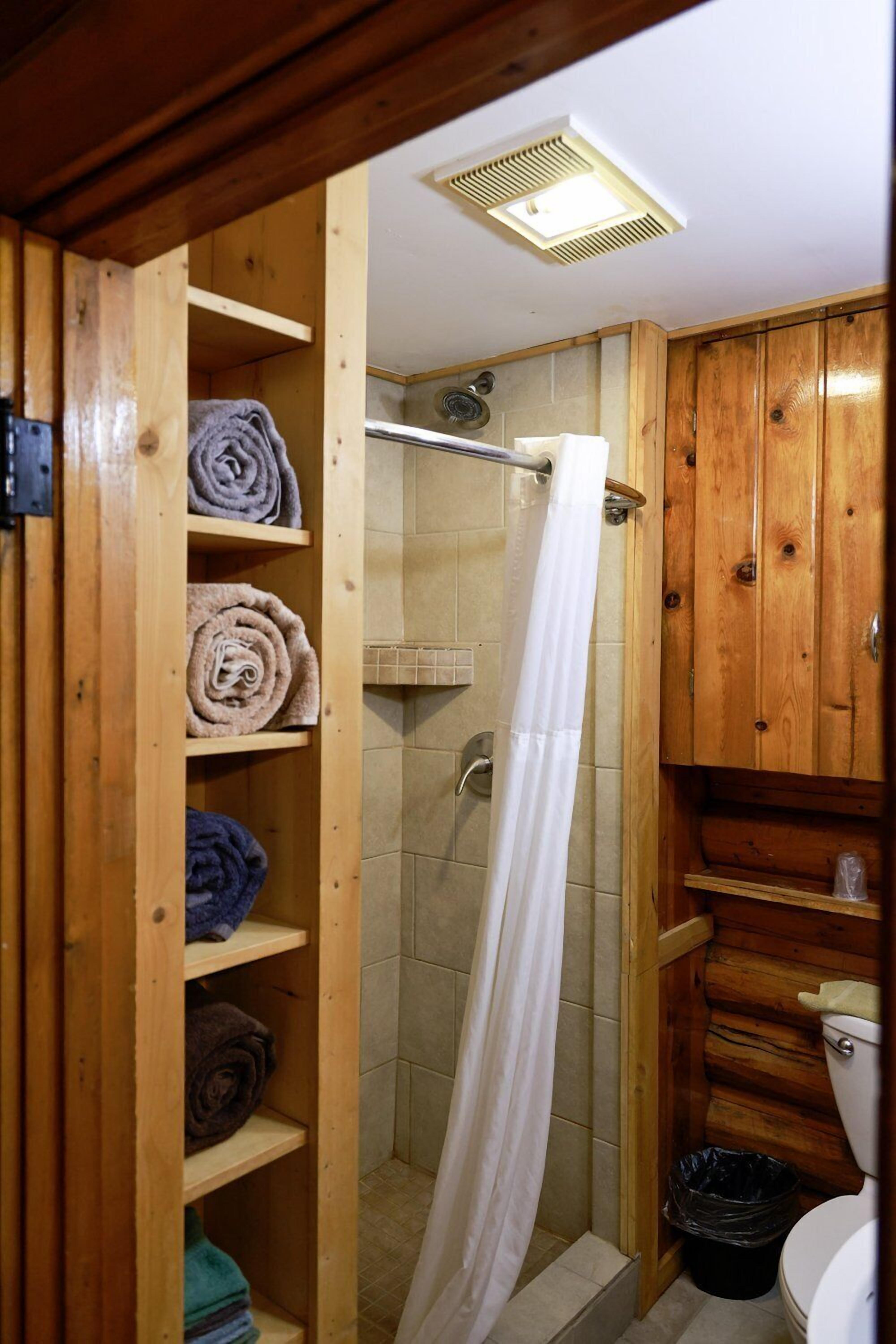 A wooden bathroom with a shower, a toilet, and shelves with towels.