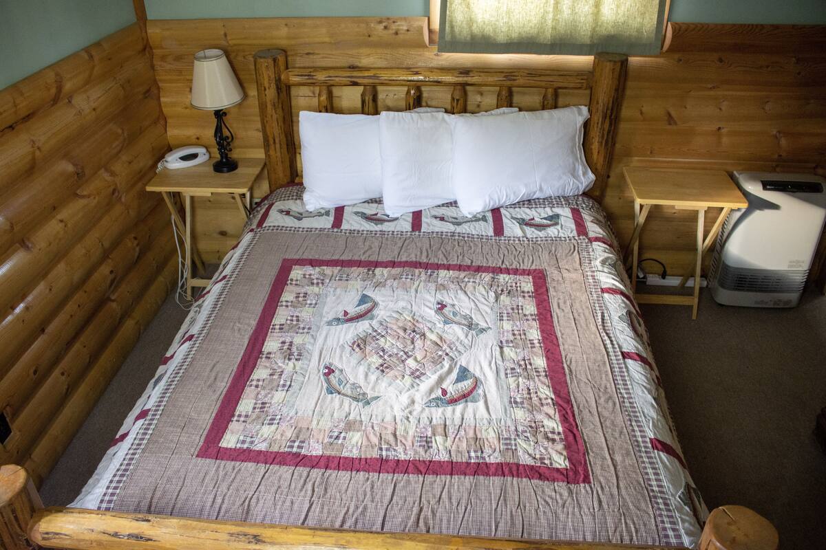 Une pièce aménagée dans le style d’un chalet en rondins de bois, avec un lit, une table de chevet dotée d’une lampe, un radiateur et une petite table.