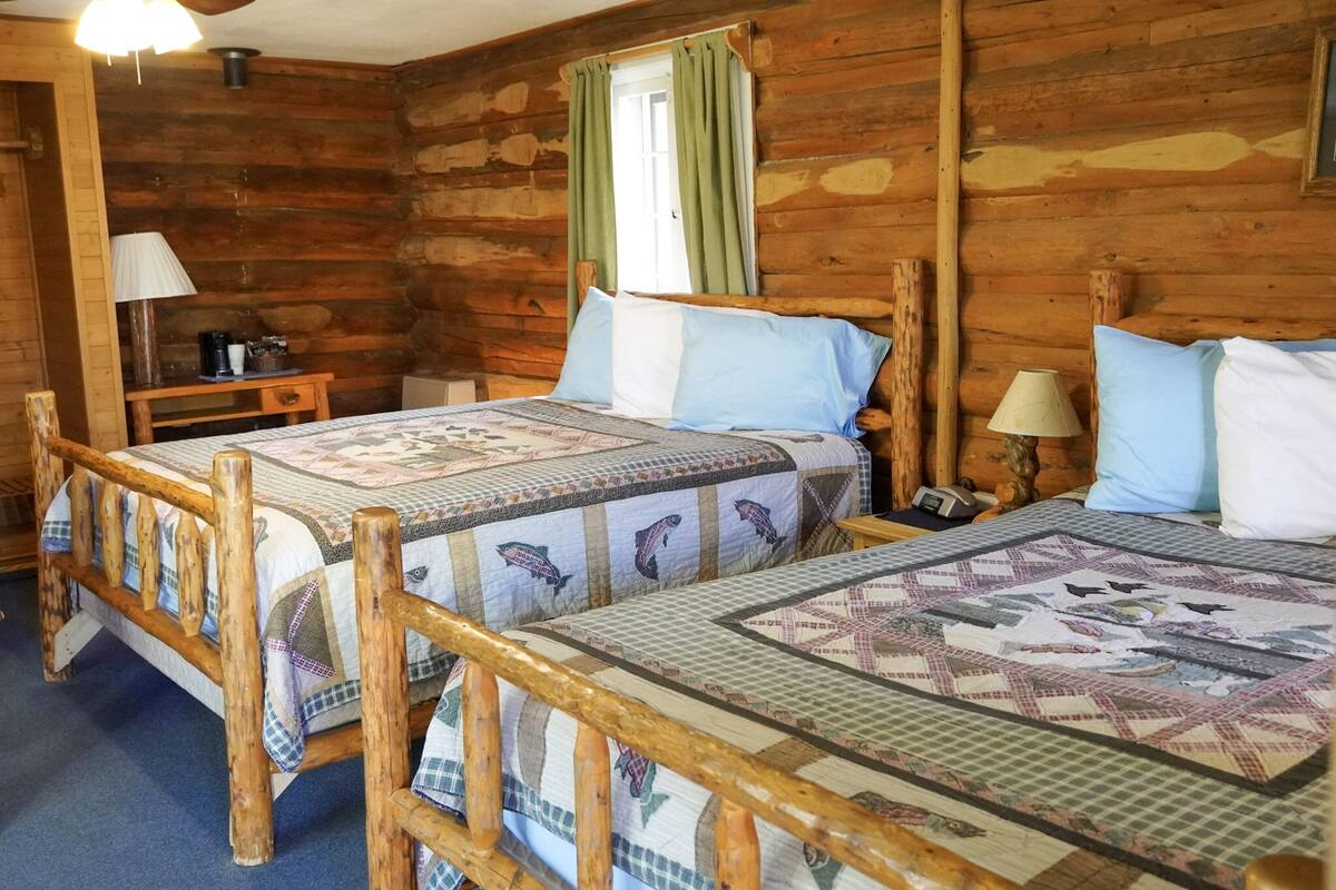 Une chambre dans un chalet en bois, avec deux lits, une table de chevet, une lampe et une fenêtre avec des rideaux.