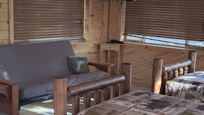 Interior acogedor de una cabaña con techo de madera, ventilador de techo, televisor montado en la pared, cama con colcha estampada y ventana con persianas.