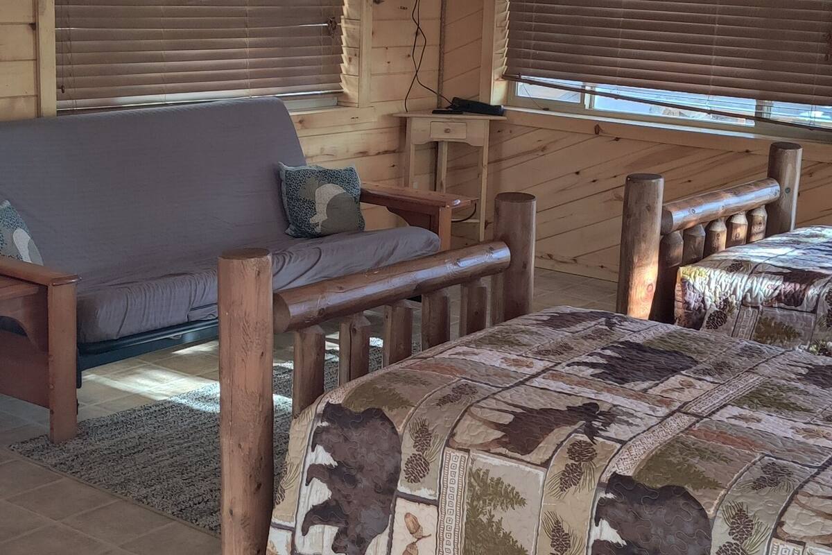 L’intérieur chaleureux d’une cabane avec un plafond en bois, un ventilateur de plafond, un téléviseur fixé au mur, un lit recouvert d’une courtepointe à motifs et une fenêtre avec des stores.