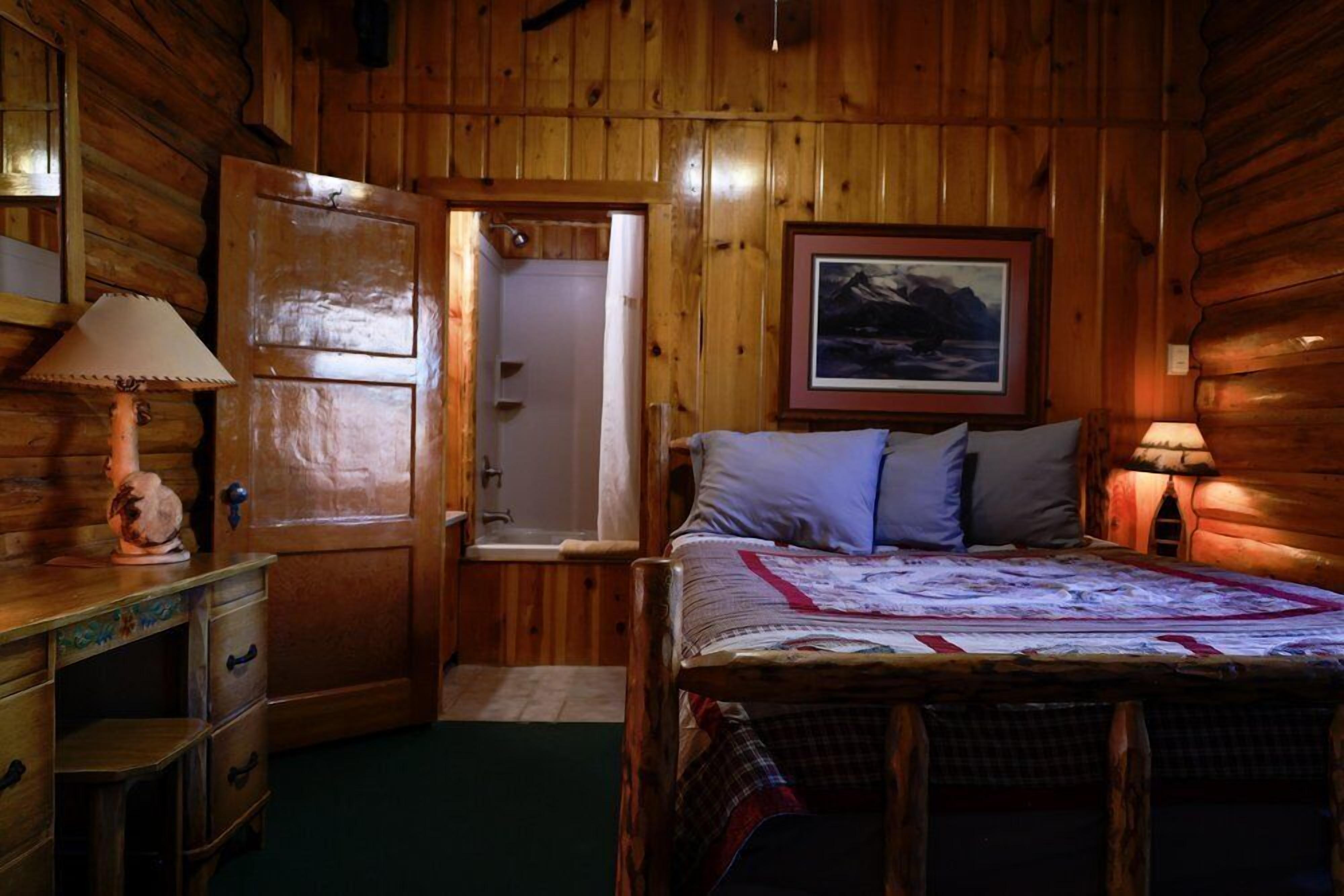 A wooden cabin room with a bed, a door, a dresser, and a framed picture on the wall.