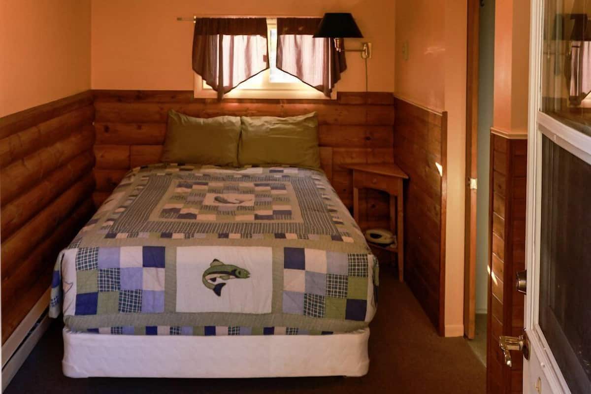 Une chambre à coucher avec une tête de lit en bois, un lit recouvert d’une courtepointe, un ventilateur de plafond, une table de chevet et une porte avec un moustiquaire.