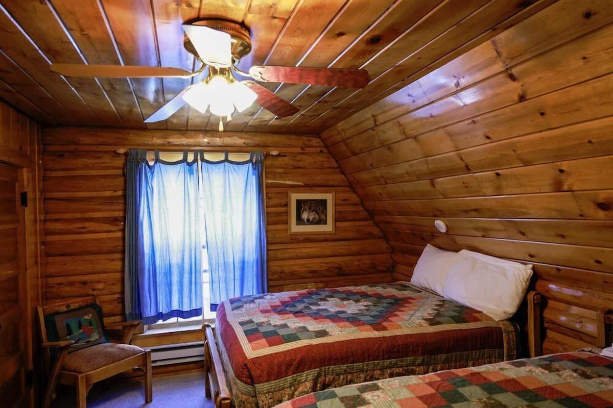Une chambre confortable dans un chalet en rondins, avec un plafond en bois, un lit recouvert d’une courtepointe, une chaise et une fenêtre avec des rideaux.