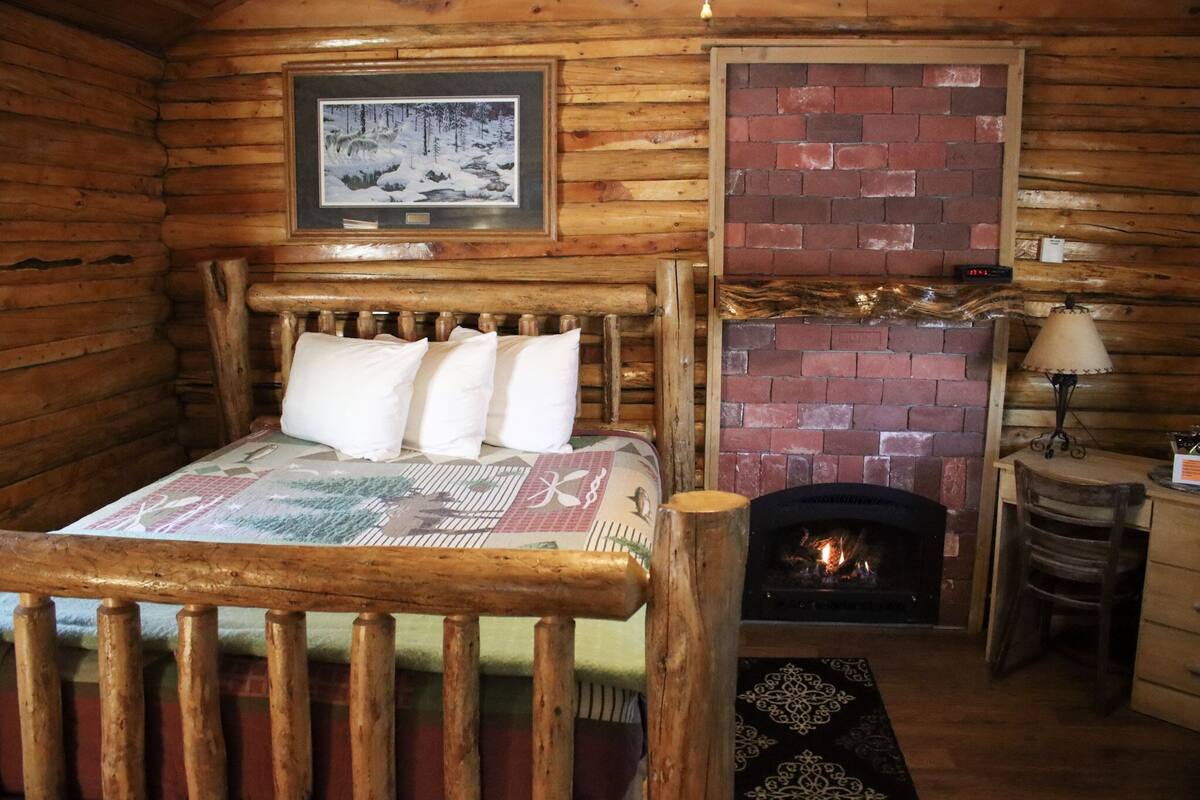 Une pièce de style cabane en bois, avec un lit, une cheminée, un tableau encadré au mur et une petite table avec une lampe.