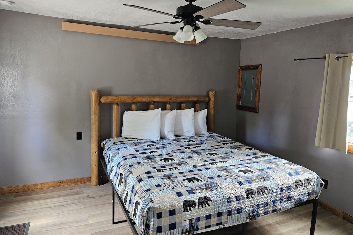 Une chambre à coucher avec une tête de lit en bois, une couvre-lit matelassée, un ventilateur de plafond et une petite fenêtre avec un rideau.