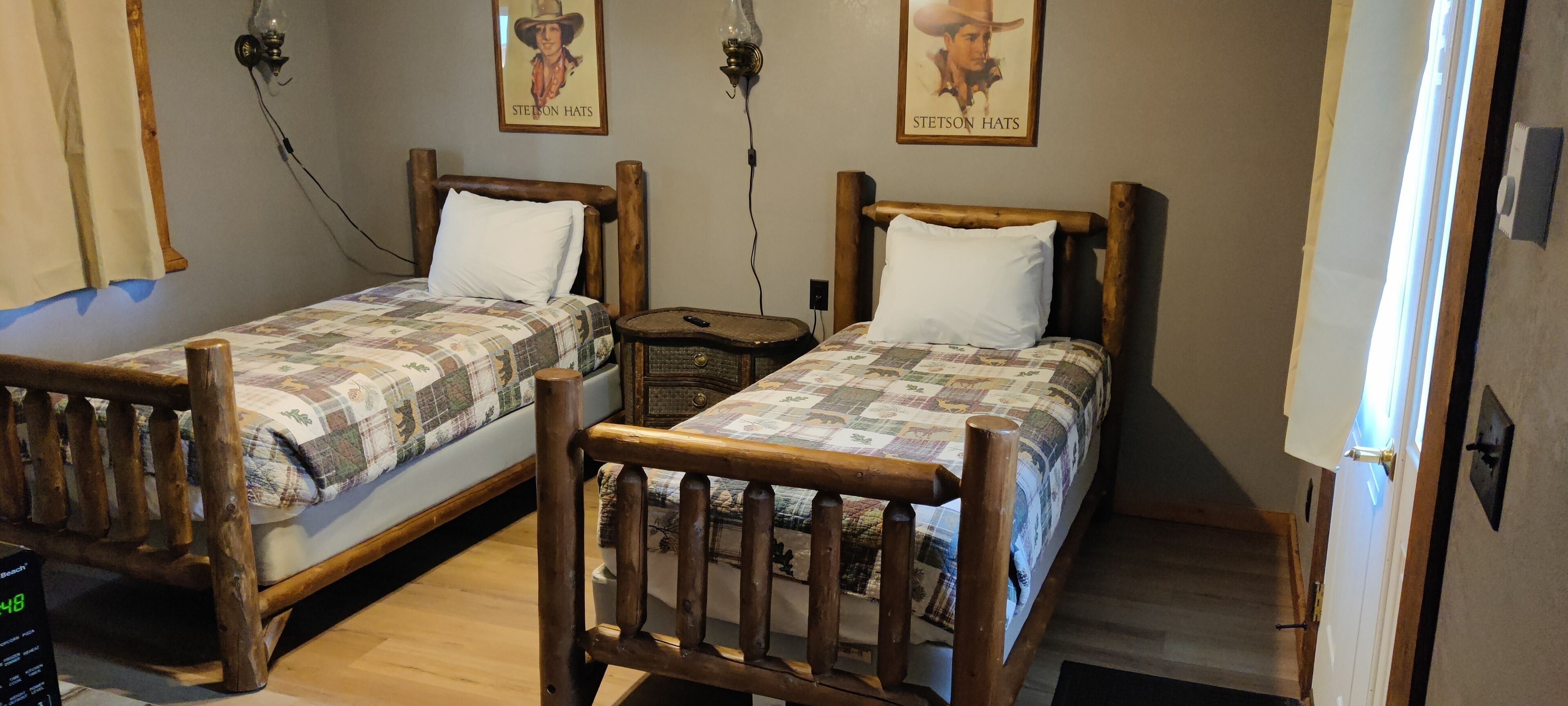 A room with two wooden beds with white pillows, a small wooden nightstand, and a wall-mounted lamp.