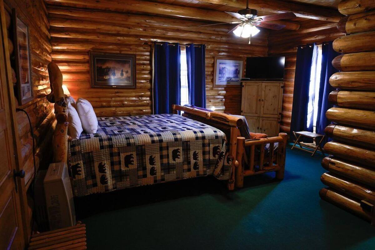 Une chambre aménagée dans un chalet en rondins, avec un lit, des meubles en bois, un ventilateur de plafond et des tableaux encadrés accrochés aux murs.