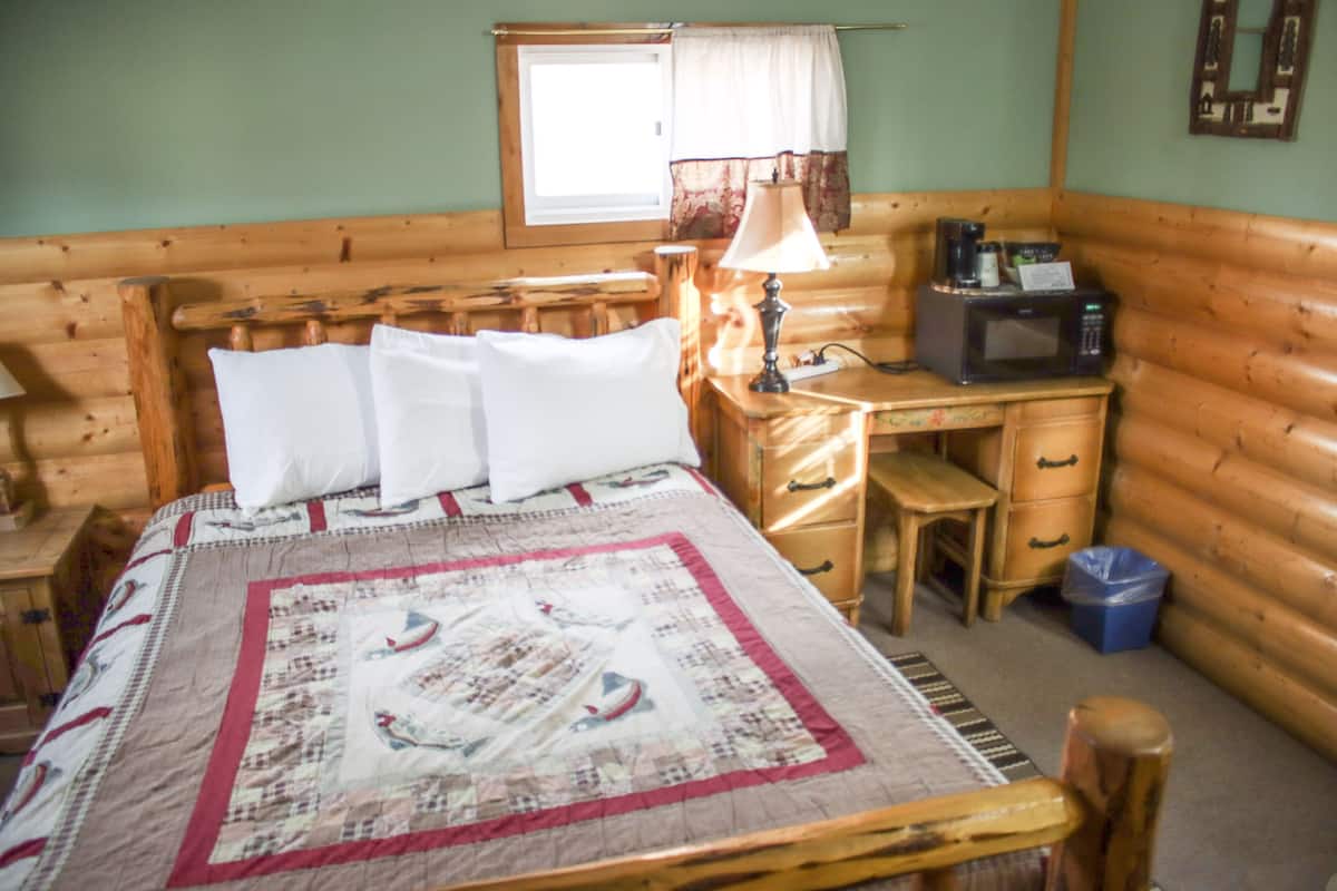 Une chambre dans une cabane en bois, comprenant un lit, un bureau, une lampe, un four à micro-ondes et une fenêtre avec des rideaux.