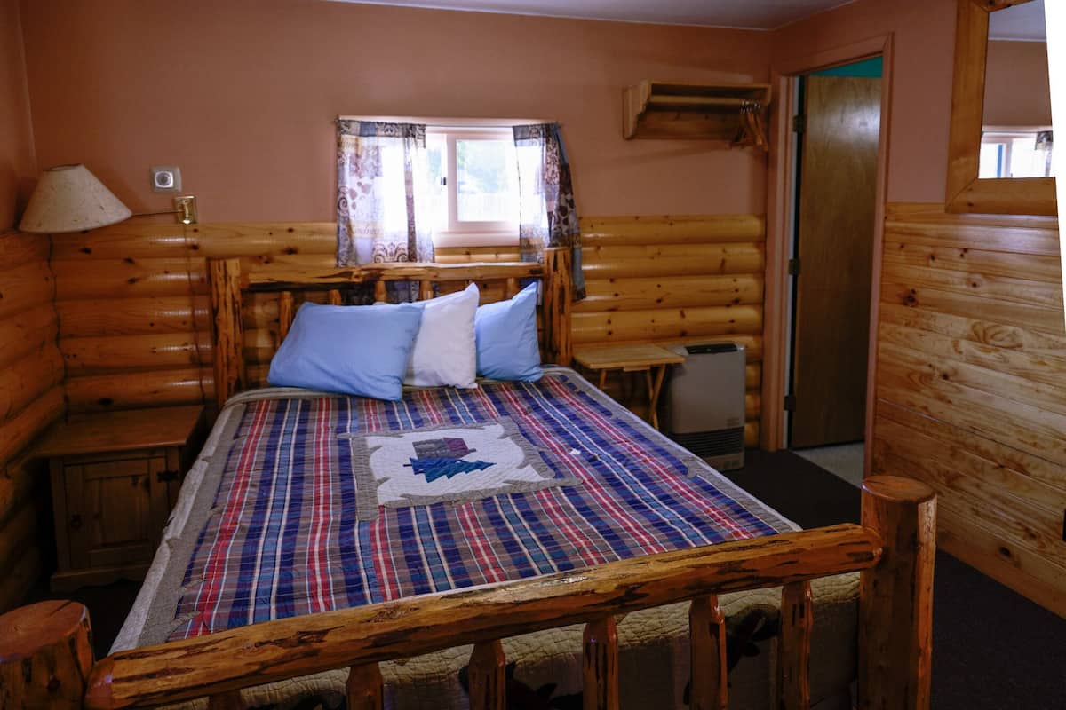 Une chambre à coucher dans un chalet en rondins, avec une couvre-lit à carreaux, une fenêtre avec des rideaux, une tête de lit en bois, une table de chevet et une lampe.
