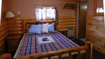 A log cabin bedroom with a plaid bedspread, a window with curtains, a wooden headboard, a bedside table, and a lamp.