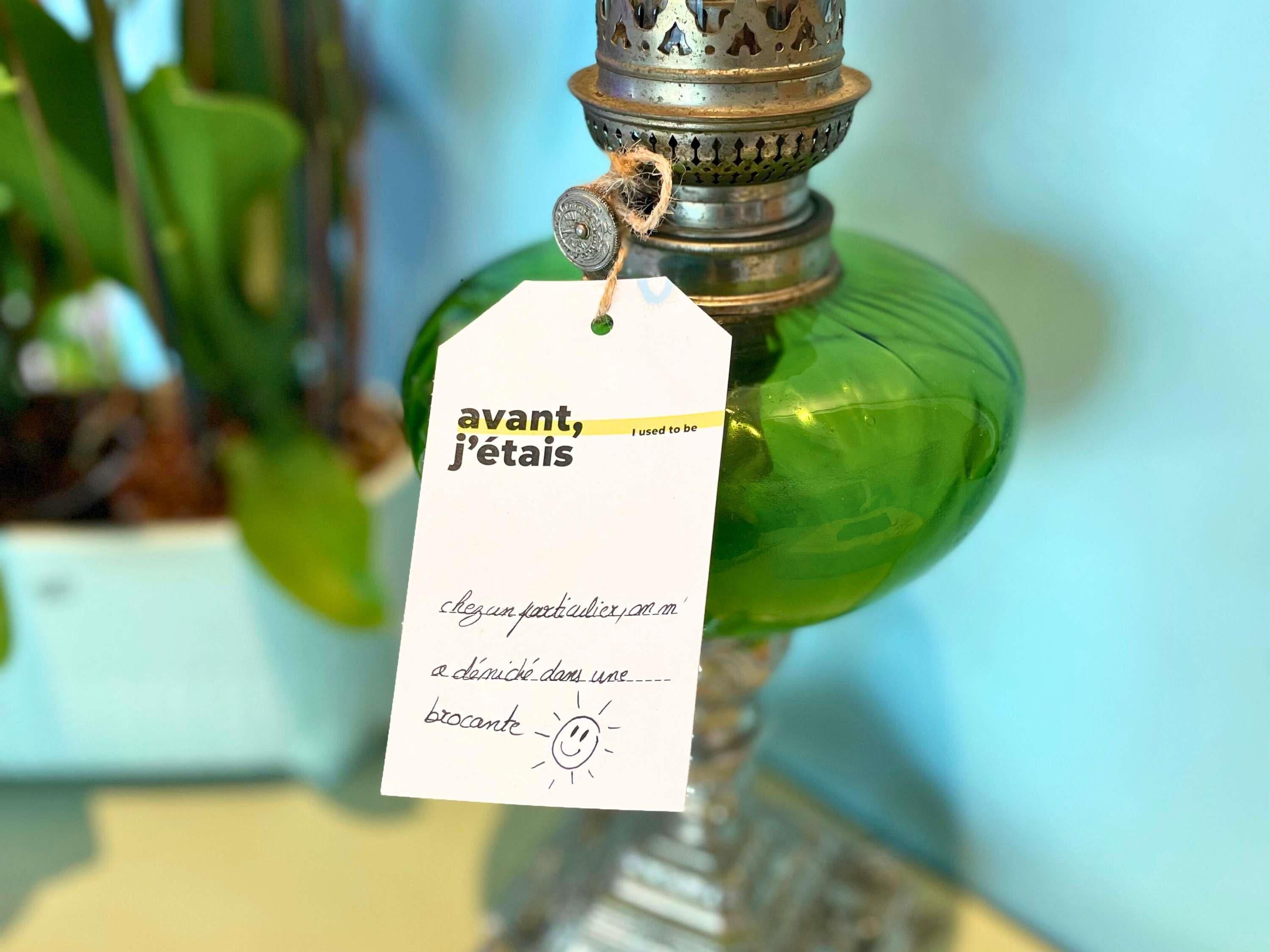 A green glass bottle with a metal top, a tag attached, and a partial view of a plant in the background.