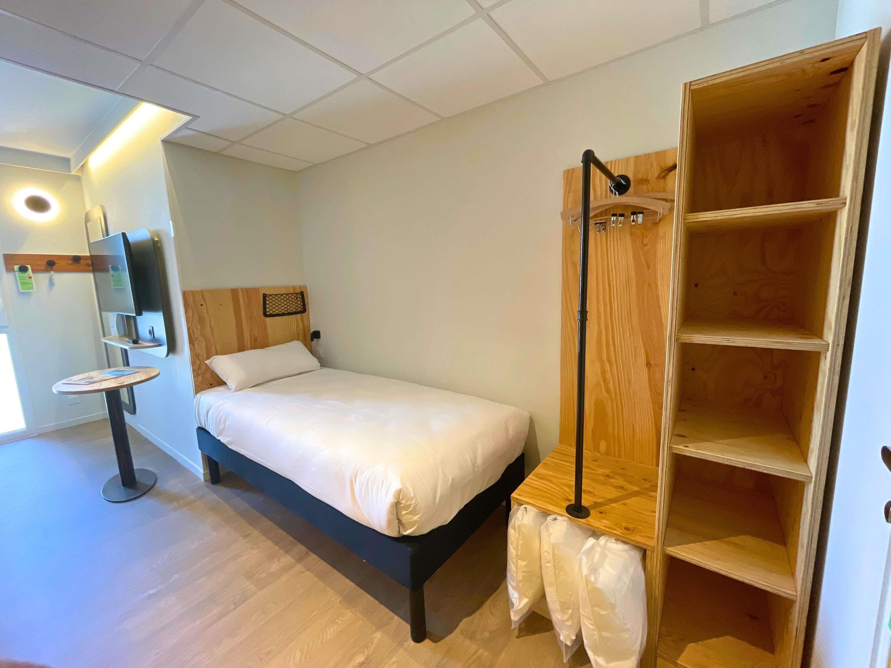 A bedroom with a bed, a small round table, a wooden wardrobe, and a built-in shelf.