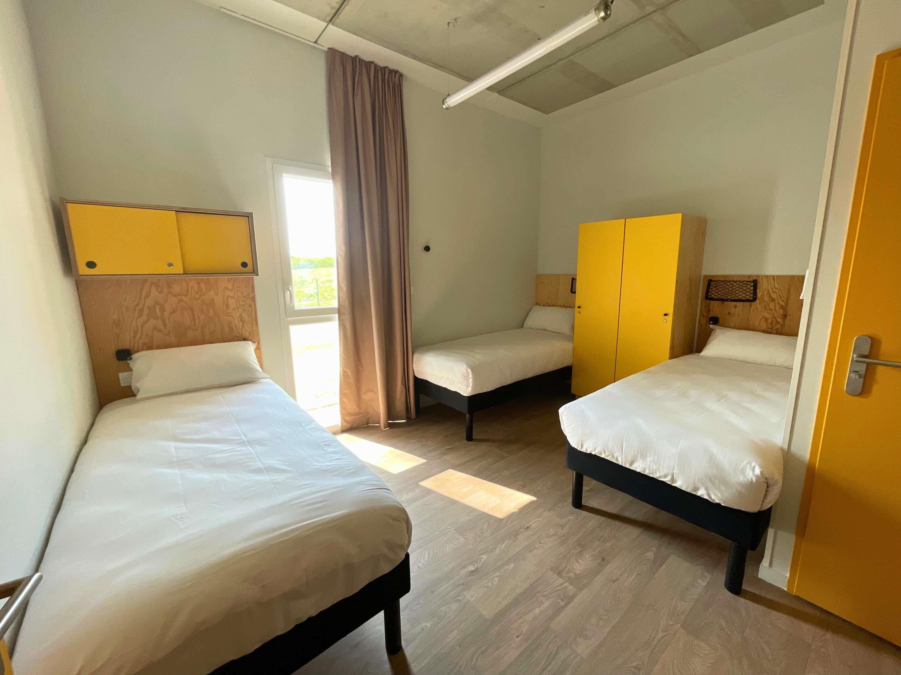 Ein Zimmer mit zwei Betten, einem Holzschrank, einer Tür und einem Fenster mit Vorhängen.