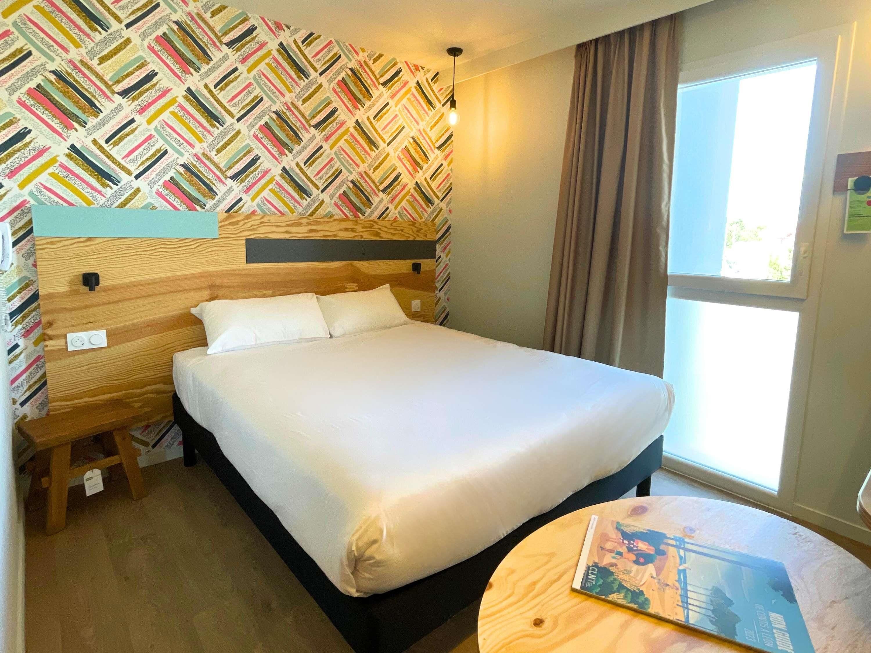 A neatly made bed with white linens in a room with a patterned wallpaper and a window with curtains.