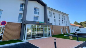 Un hôtel moderne, reconnaissable à son grand panneau vert, à son entrée en verre et à son parking où se trouve un fourgon blanc.
