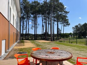 Une terrasse avec une table ronde en bois et des chaises orange, entourée de grands pins.