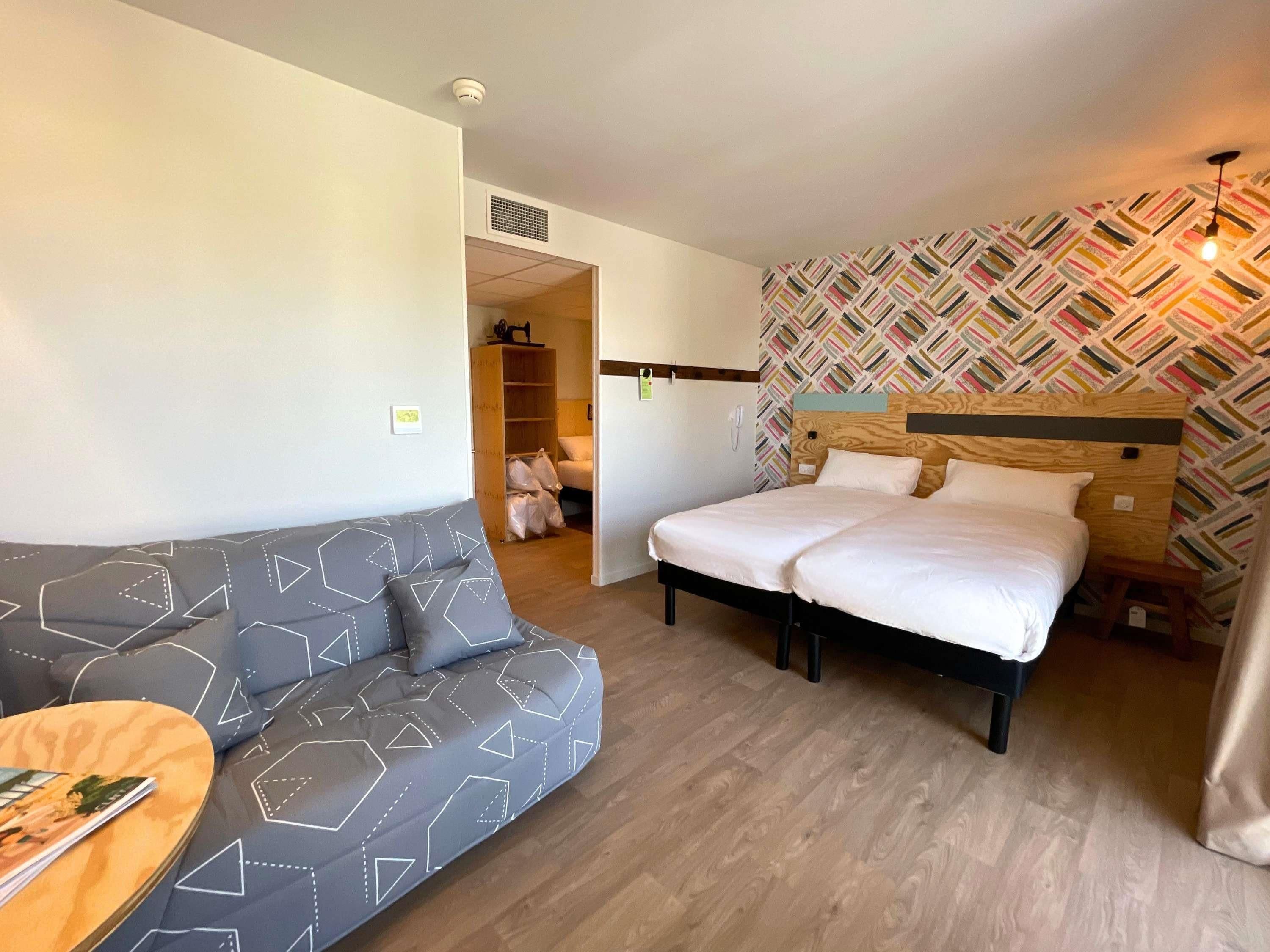 A hotel room with a bed, a sofa, a wooden floor, a patterned wall, and a ceiling light.